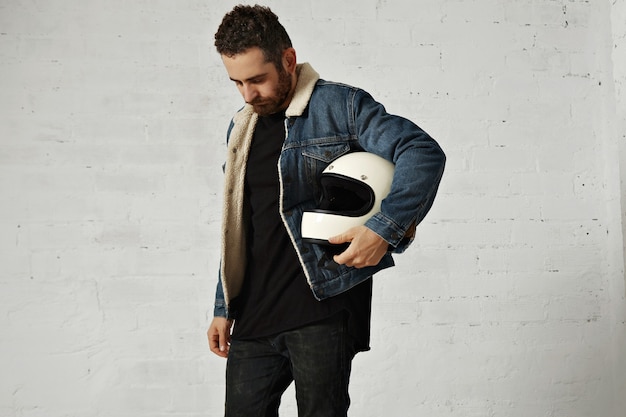 Free Photo motor biker wears shearling denim jacket and black blank henley shirt, holds vintage beige motorcycle helmet, looking down, isolated in center of white brick wall