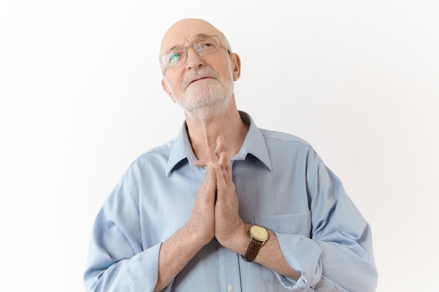 Free photo mournful old mature man in eyeglasses and blue shirt having hopeful facial expression, keeping hands pressed together in prayer, hoping for the best while facing difficulties, stress or problems