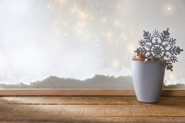 Free Photo mug with toy snowflake on wood table near bank of snow and fairy lights