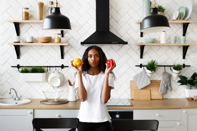 Mulatto woman dressed in white t-shirt, with funny face and loose hair is holding red and yellow peppers in hands near cheeks on the modern kitchen