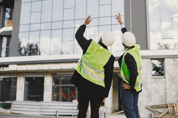 Multi racial builders standing outdoors, back view. Wearing uniform, talking about new glass building. Working on the poject. City infrastructure