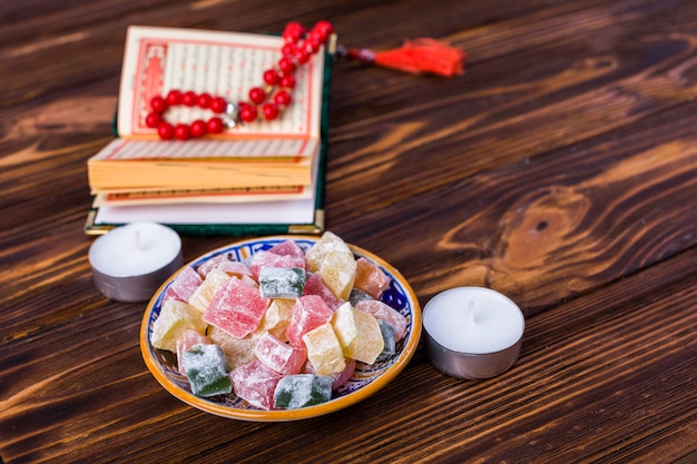 Free photo multicolor cubes of rakhat-lukum in plate with islamic holy book; rosary beads and candles on wooden desk