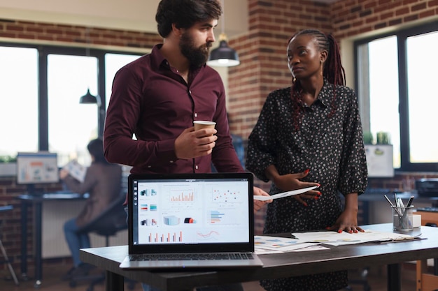 Multiethnic company employees reviewing annual company revenues and discussing startup marketing strategy. Financial agency coworkers brainstorming project ideas with laptop showing charts on display.