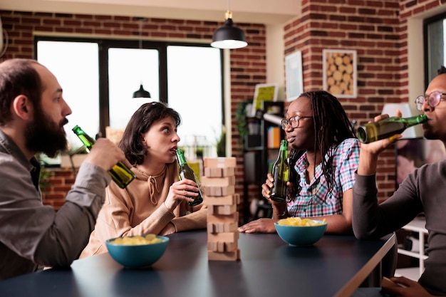 Free photo multiethnic friends looking at each other while sipping beer and playing with wood blocks. diverse people sitting together at home in living room while talking and enjoying fun leisure activity.