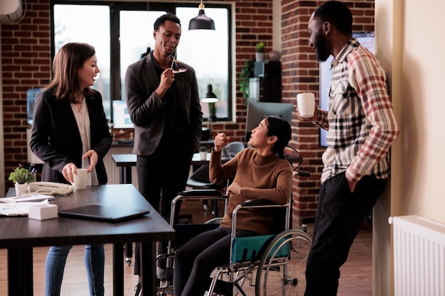 Multiethnic group of colleagues laughing in business office with paralyzed woman sitting in wheelchair. Asian employee with chronic disability and impairment having fun with coworkers.