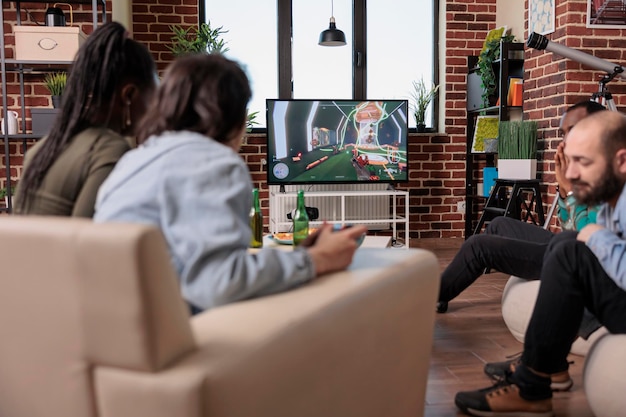 Multiethnic group of friends having fun with shooter competition, playing shooting video games on tv console at home with friends. Enjoying game play at gathering, drinking beer.