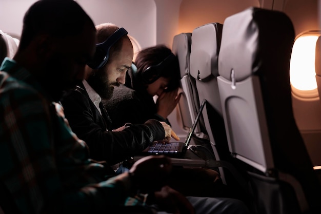 Multiethnic group of people flying in economy class together to arrive at destination, man using laptop computer during international flight. Tourists travelling abroad by airplane.