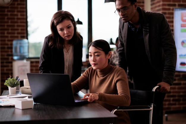 Multiethnic team of partners working with paralyzed woman sitting in wheelchair, suffering from chronic disability and impairment. Business people using laptop, health condition.