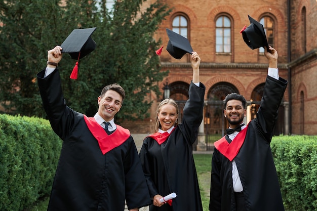 Multinational graduates male and female celebrating graduation in university campus, removing their graduation hats and smiling to the camera.