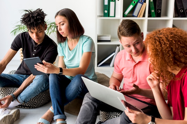 Free photo multiracial friends using electronic gadgets sitting on floor having discussion