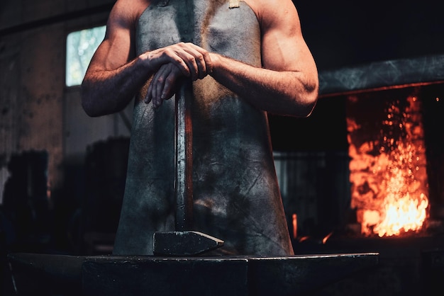 Muscular blacksmith in protective apron at his workshop with hammer in his hands. There are fire at background.