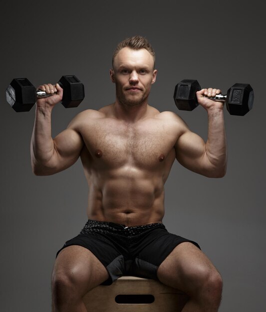 Muscular bodybuilder guy sitting on chair and doing exercises with dumbell on grey background