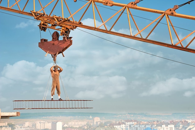 Muscular builder with bare torso standing on iron construction on high and holding by ropes. Man wearing hat and work wear looking at camera. Blue sky with clouds on background.