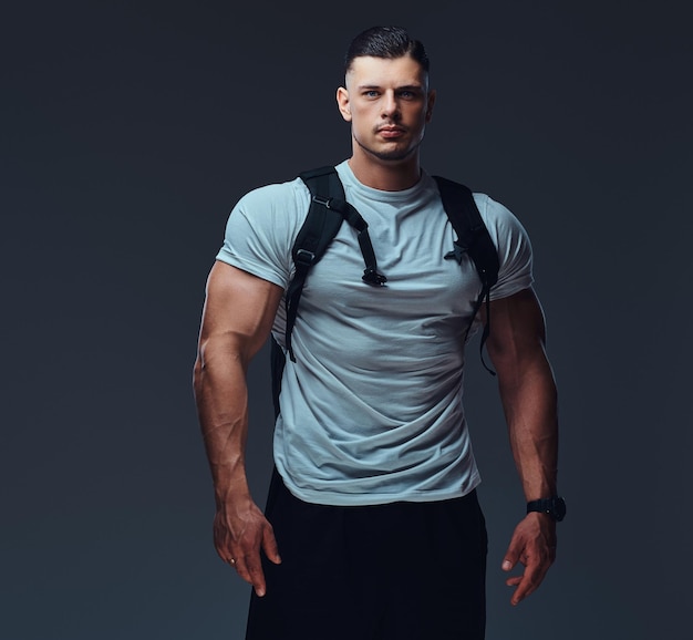 Free photo muscular handsome bodybuilder in sportswear with rucksack posing against a gray background.