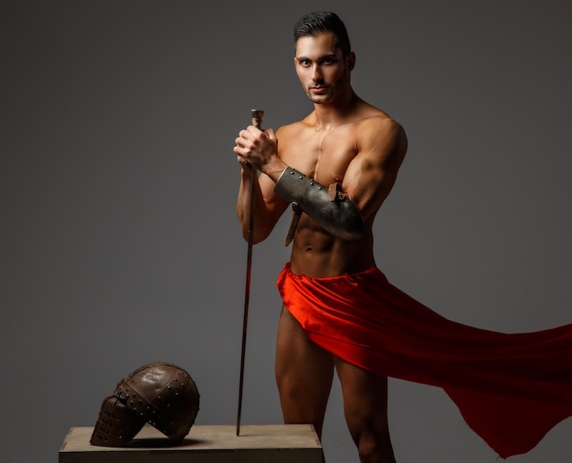 Free photo muscular man ancient rome soldier holding a sword dressed in a red fluttering dress.