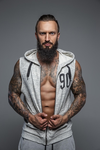 Muscular tattooed man with beard in grey zipuo hoodies. Isolated on grey background.