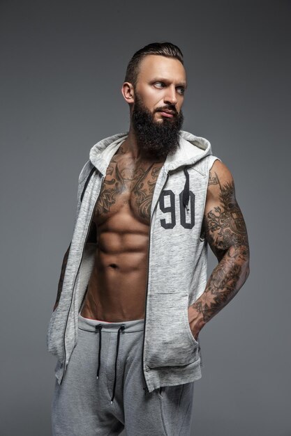 Muscular tattooed man with beard in grey zipuo hoodies. Isolated on grey background.