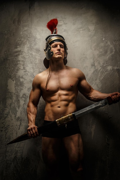 Muscular warrior with sword and helmet posing in front of concrete wall