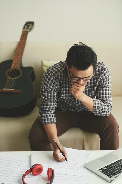 Free photo musician sitting on couch and writing song, and laptop, headphones and guitar lying around