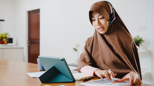 muslim lady wear headphone using digital tablet talk to colleagues about sale report in conference video call while working from home at kitchen.