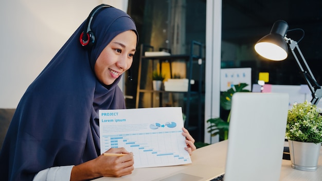 muslim lady wear headphone using laptop talk to colleagues about sale report in conference video call while working from home office at night.