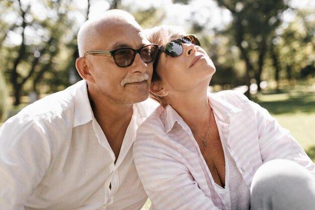 Mustachioed man in black sunglasses and white cool outfit posing with blonde smiling woman in pink striped clothes outdoor