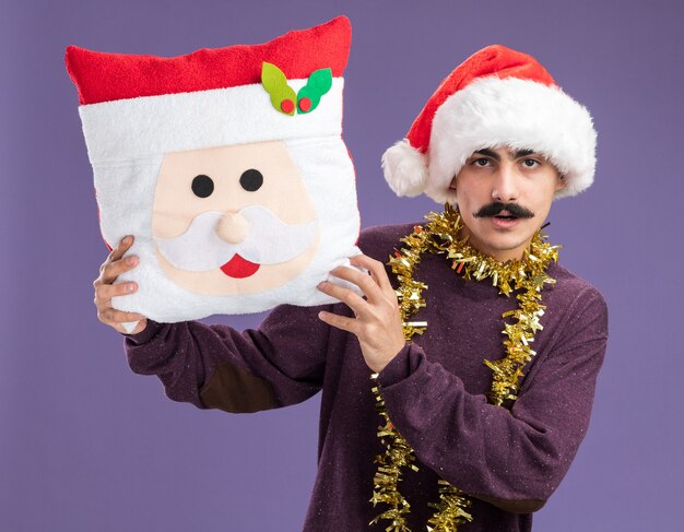 Mustachioed man wearing christmas santa hat  with tinsel around his neck holding christmas pillow looking at camera with serious face standing over purple background