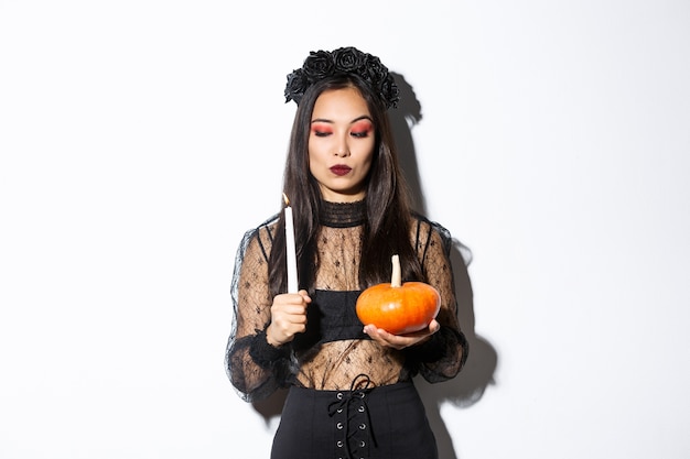 Free photo myserious asian wicked witch in gothic dress, looking at lit candle, holding pumpkin, standing over white background.