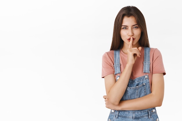 Mysterious alluring modern brunette woman in overalls tshirt prepare surprise asking keep quiet about her secret shushing say shhh as press index finger to lips squinting sly white background