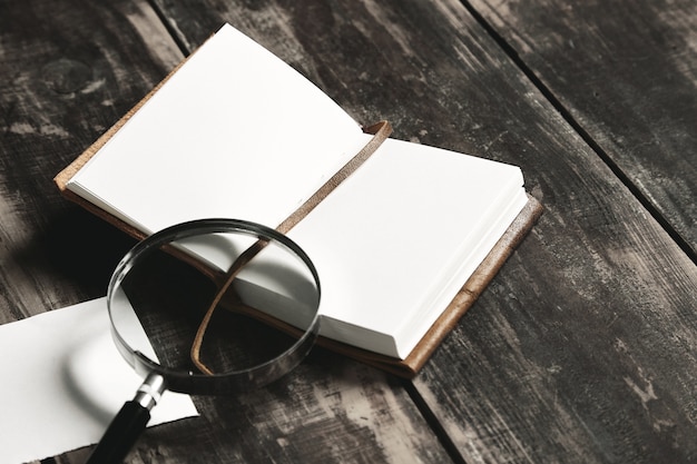 Free photo mysterious detective game concept. opened notebook in leather cover, sheet of white paper and big vintage magnifier isolated on black aged wood table, closeup, side view