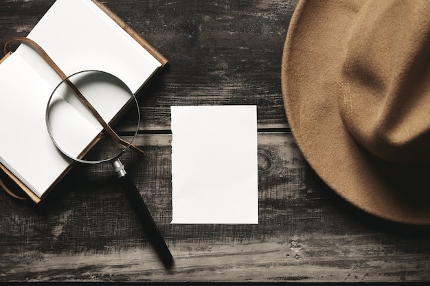 Free Photo mysterious detective game concept. opened notebook in leather cover, sheet of white paper, felt brown hat and big vintage magnifier steel glasses isolated on black aged wood table. top view.