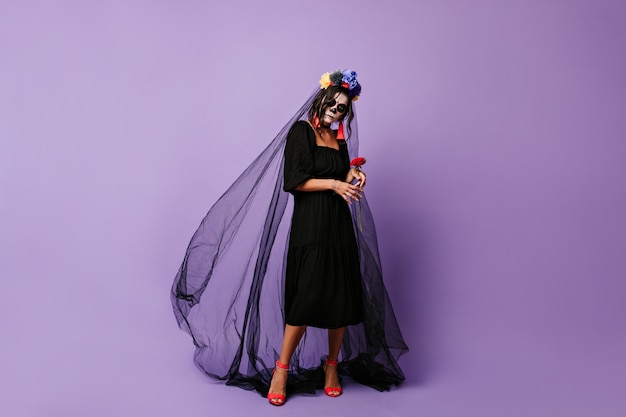 Mysterious woman gently holds red rose. girl with makeup for Halloween in black dress and veil.