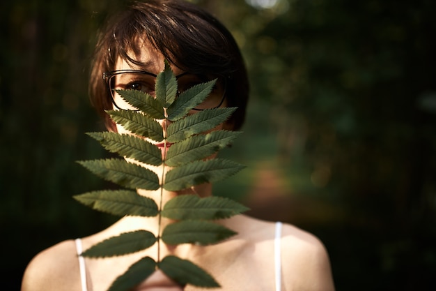 Mysterious young woman with short brunette hair and dark eyes posing in woods, covering face with large green leaf, enjoying wild nature