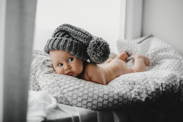 Naked newborn boy in a warm winter hat lies on the soft blanket before a bright window