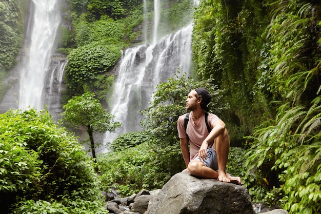 Free Photo nature, tourism and people. young barefooted tourist wearing jeans short and backpack sitting on big rock deep inside green rainforest