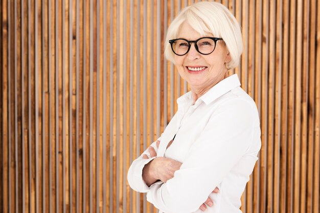 Neat old woman with eyeglasses standing next to a wooden wall