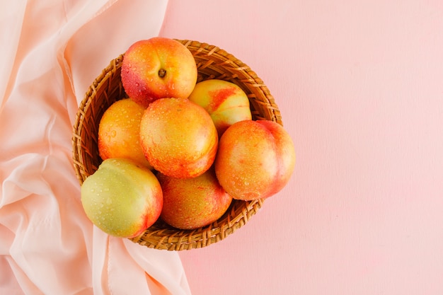 Free photo nectarines in a basket on pink and textile surface, flat lay.