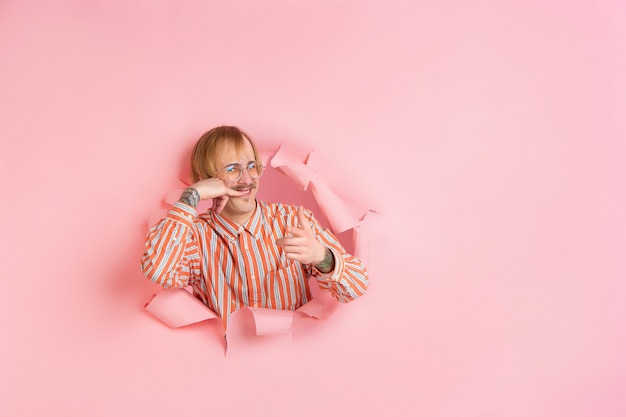 Free photo need you to call me. cheerful caucasian young man poses in torn coral paper, emotional expressive.