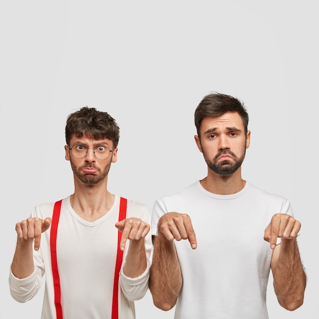 Free photo negative facial expressions and feeling concept. upset two men with gloomy expressions point down, frown faces with displeasure, dislike something, pose over white wall with blank space