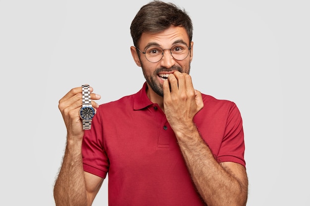 Free photo nervous bearded male bites finger nails, holds wrist watch, worries as being late for important meeting, dressed in casual t-shirt. embarrassed young man waits for something. times flies quickly