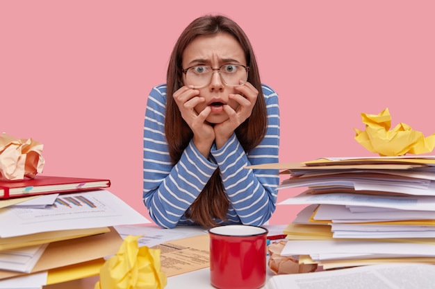 Free photo nervous displeased lady stares directly at camera keeps hands under chin, wears blue striped jumper, square glasses, afraids of deadline