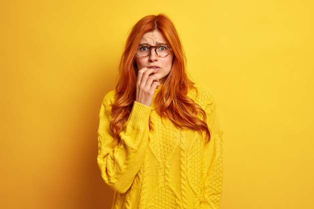 Nervous redhead young woman looks worried feels embarrassed frowns face wears optical glasses and sweater.