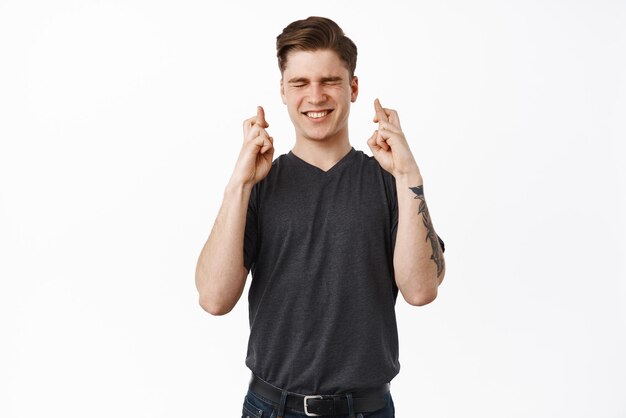 Nervous young man cross fingers for good luck and waiting for wish come true praying anticipating news pleading for good exam mark white background