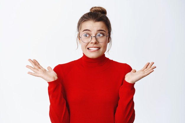 Free photo nervous young woman in glasses worried as making mistake, shrugging and looking aside indecisive, know nothing, cant understand, white wall