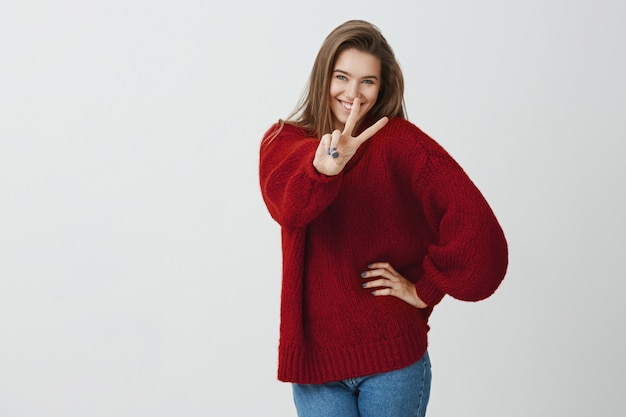 Never stop smiling. Studio shot of positive beautiful caucasian woman pulling hand towards camera with v sign, showing peace gesture, expressing friendliness and joy, standing 