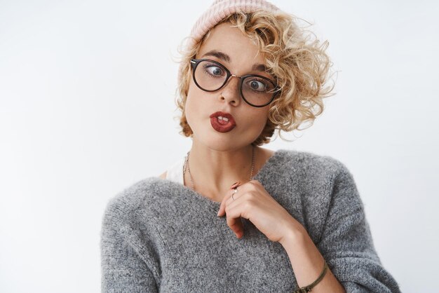 Free photo never too old for fooling around. portrait of playful and immature funny cute blond girlfriend in beanie and winter sweater squinting folding lips and showing tongue aping on white wall. copy space