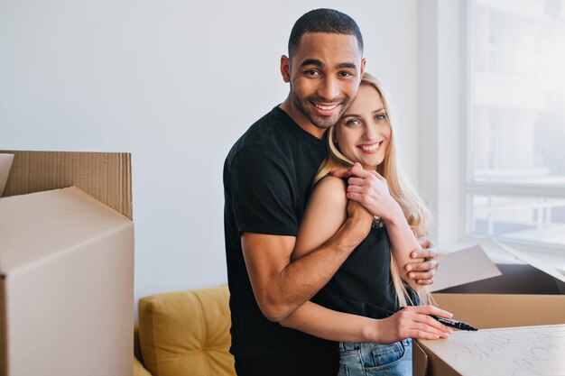 New family going to move in new apartment, joyful couple packing for relocation. Young man and woman hugging, they around boxes in empty room. Wearing casual clothes.