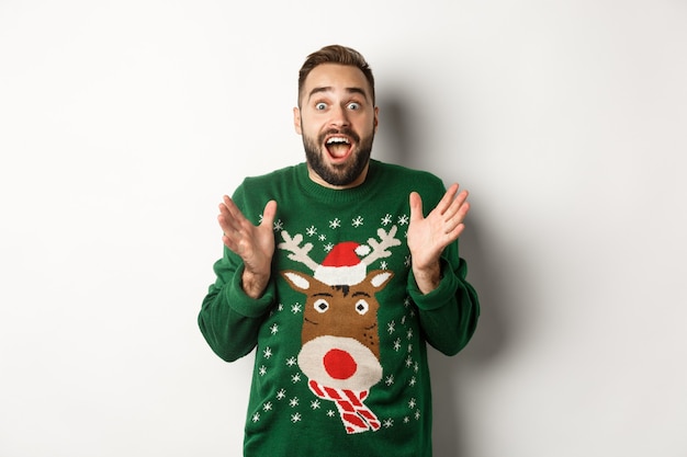 Free photo new year celebration and winter holidays concept. happy and surprised bearded guy looking amazed, catching something, standing in funny christmas sweater, white background.