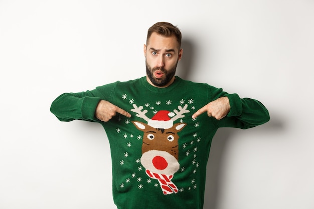 Free photo new year celebration and winter holidays concept. man pointing at his funny christmas sweater and looking confused, standing over white background.