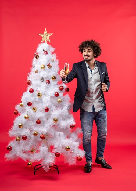 New year celebration with bearded young man holding wine and standing near decorated white Christmas tree